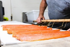 A person holding a wooden rack with several orange-hued salmon fillets, likely caught from the rich waters of Skeena, lays them out on a white surface for drying or smoking. The person is partially visible, wearing a grey shirt. The background features kitchen equipment reminiscent of Kitimats fishing culture. Skeena Adventure Fishing